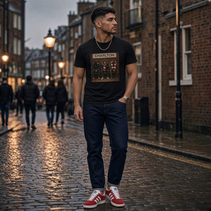 A man stands on a wet cobblestone street wearing a black Charlton graphic t-shirt, dark navy jeans, and red low-top sneakers. The background shows a blurred London street scene with brick buildings and streetlights.