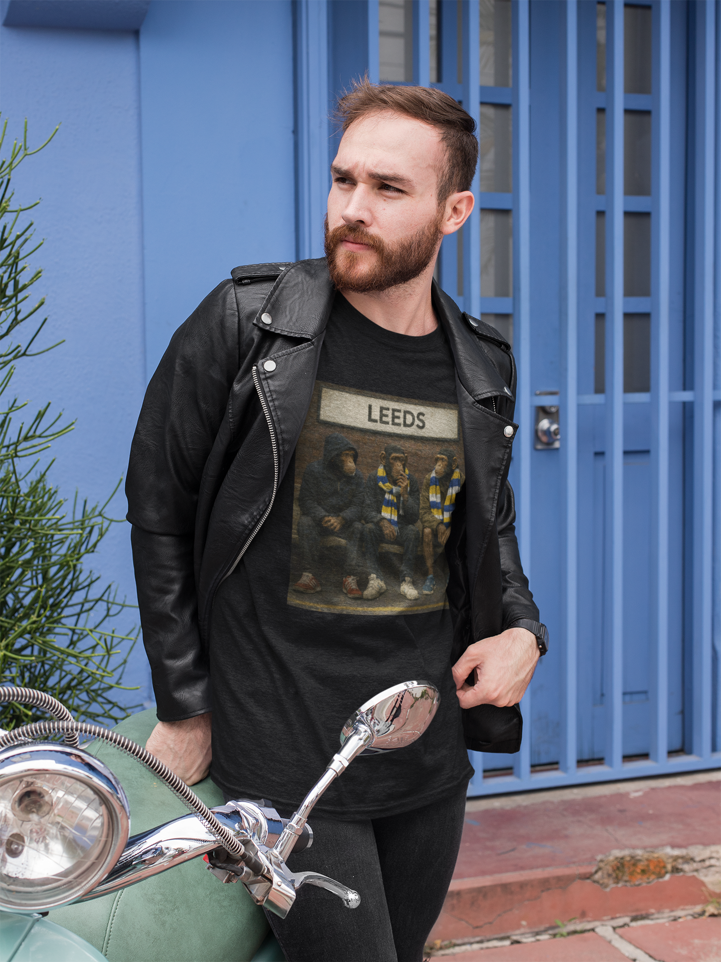 A man wearing a black leather jacket over a black graphic Leeds t-shirt with 'LEEDS' printed on the chest, standing next to a green scooter in front of a blue door.