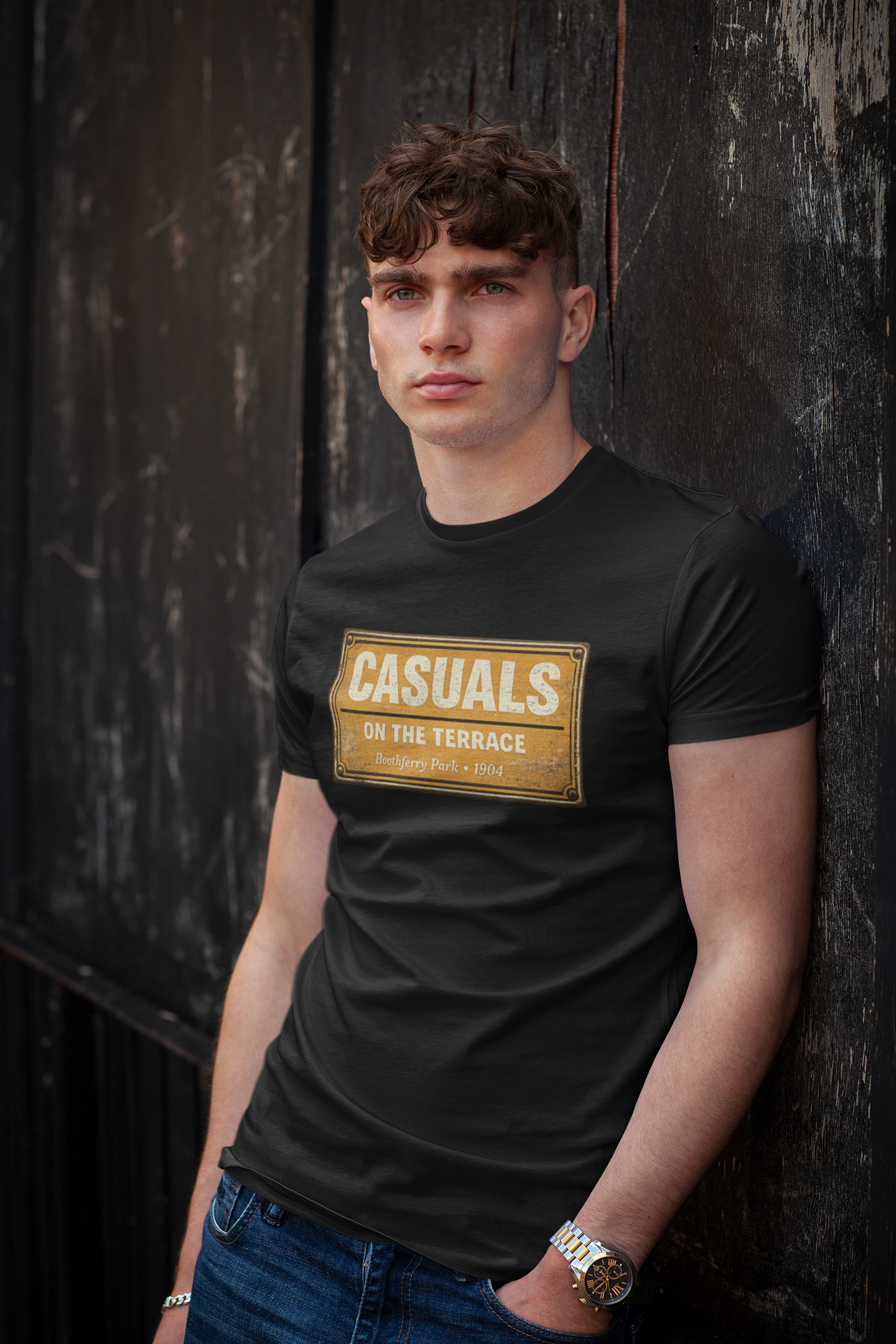 A man wearing a black HULL CITY  t-shirt with a gold graphic print that reads 'CASUALS ON THE TERRACE', leaning against a dark wooden wall.