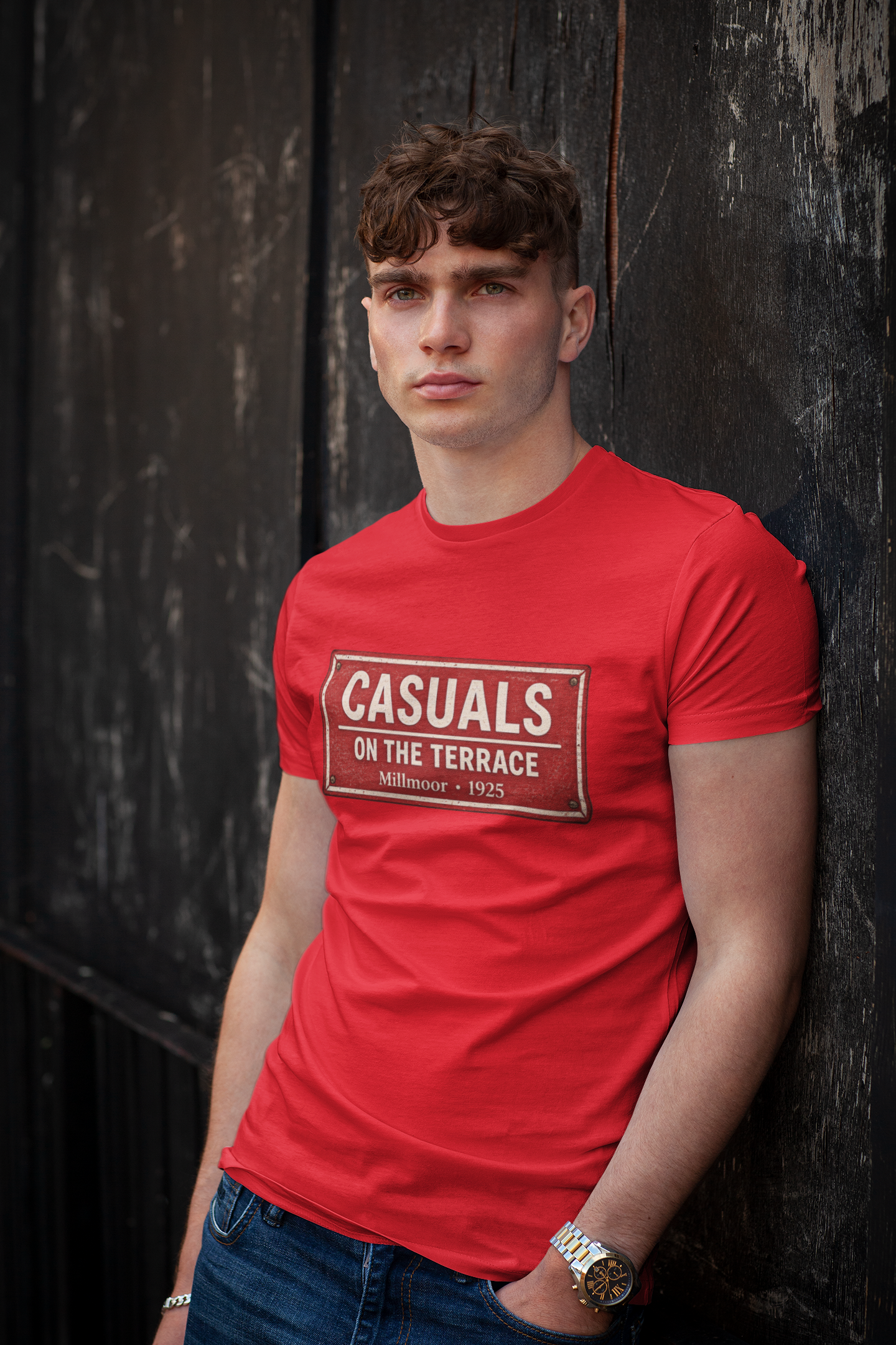A young man with curly brown hair wearing a bright red Rotherham t-shirt with a vintage-style graphic that reads 'CASUALS ON THE TERRACE' in white text on a brown background. He is leaning against a dark, weathered wooden wall, wearing blue jeans and a gold watch.