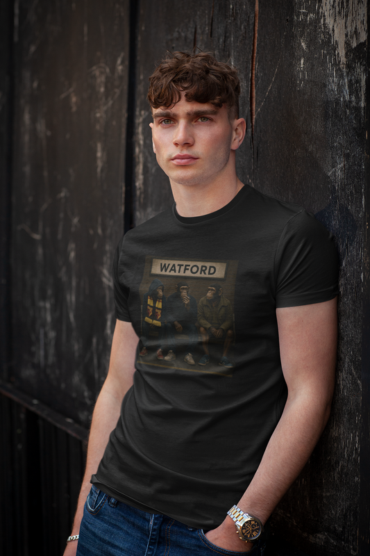 A young man with curly brown hair leans against a dark wooden wall, wearing a black Watford t-shirt with a distressed Watford print and blue jeans.