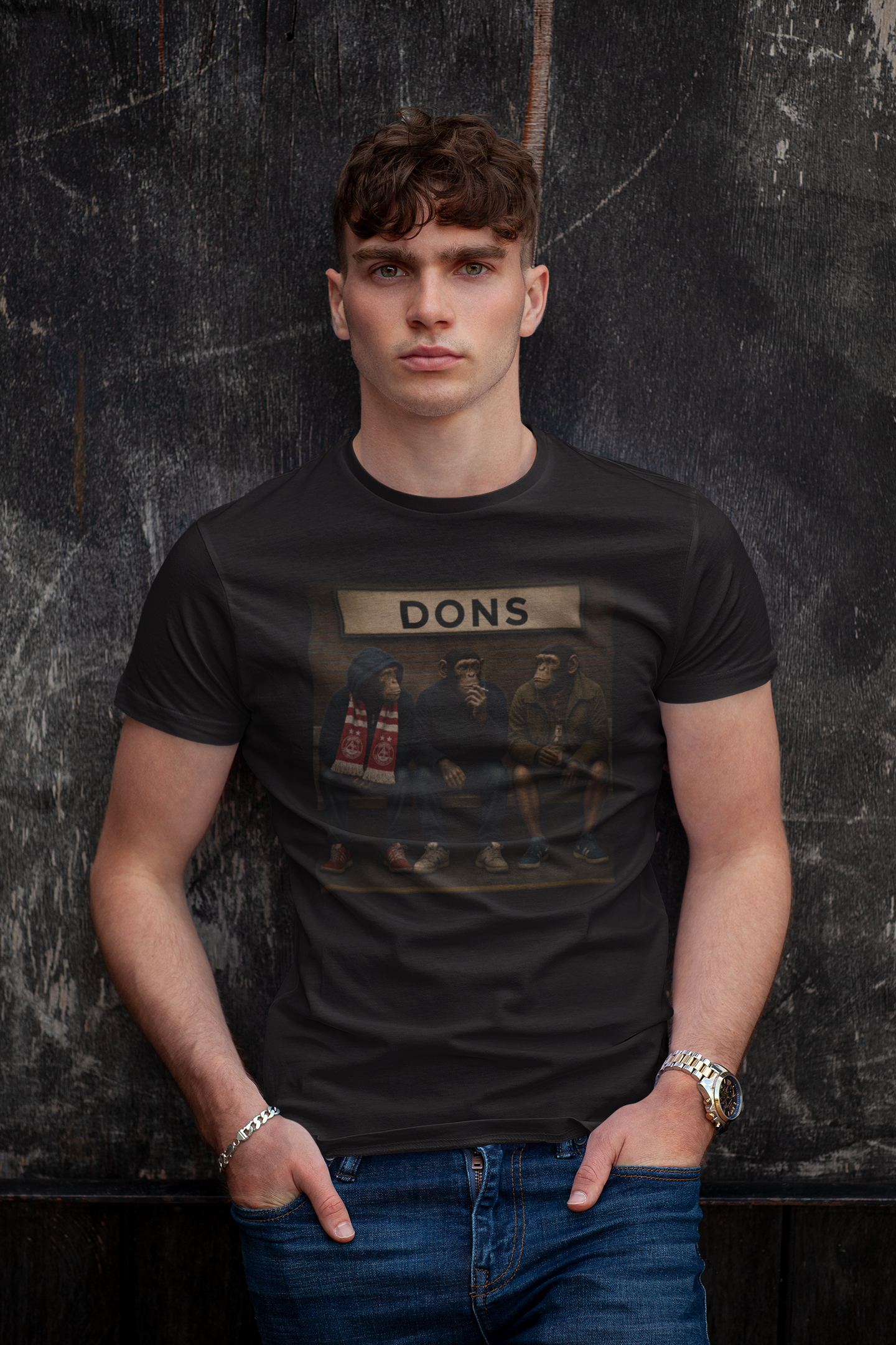 A man wearing a black graphic Aberdeen t-shirt with a vintage-style print of the band Dons and the word 'DONS' on a banner, paired with blue jeans, standing against a dark textured wall.