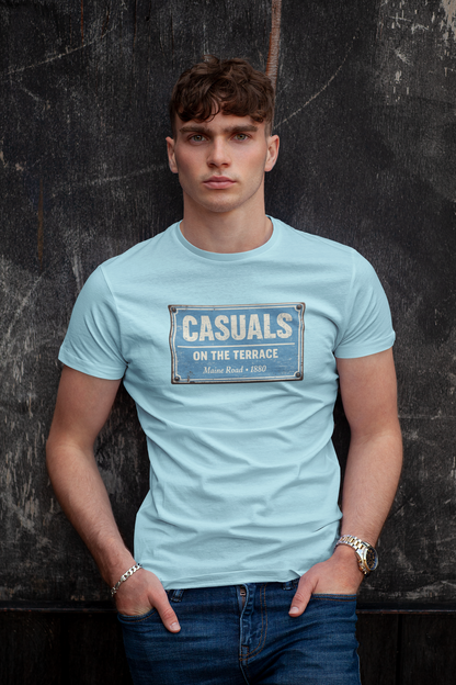 A man wearing a light blue MCFC t-shirt with a vintage-style graphic that reads 'CASUALS ON THE TERRACE' in white text on a distressed blue sign design. He is posing with his hands in his pockets against a dark, textured wall.
