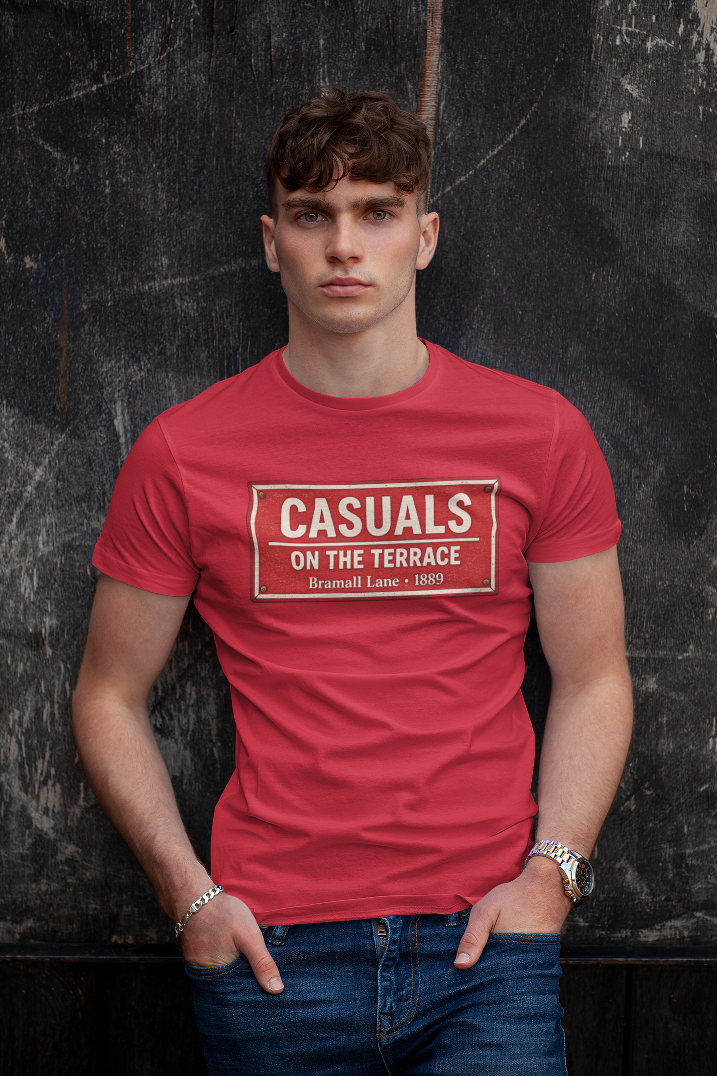 A man wearing a red SUFC t-shirt with a vintage-style street sign graphic that reads 'CASUALS ON THE TERRACE' in white, posed against a dark textured wall.
