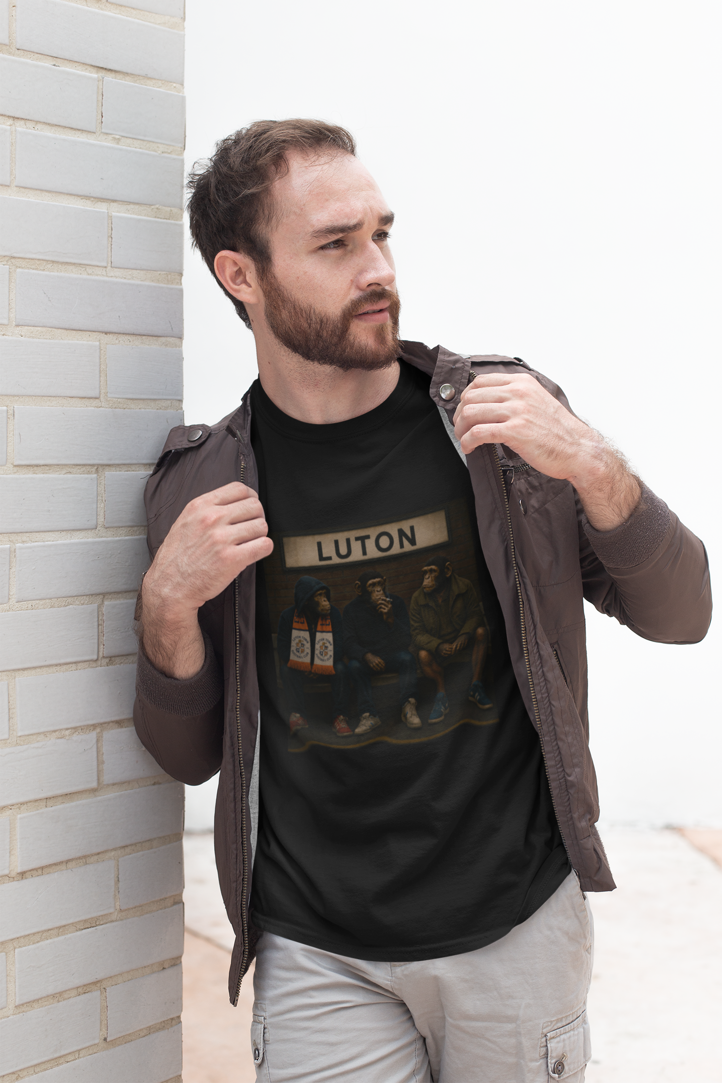 A man wearing a black graphic Luton t-shirt with 'LUTON' printed on the chest, styled with a brown jacket and light gray cargo pants, leaning against a white brick wall.
