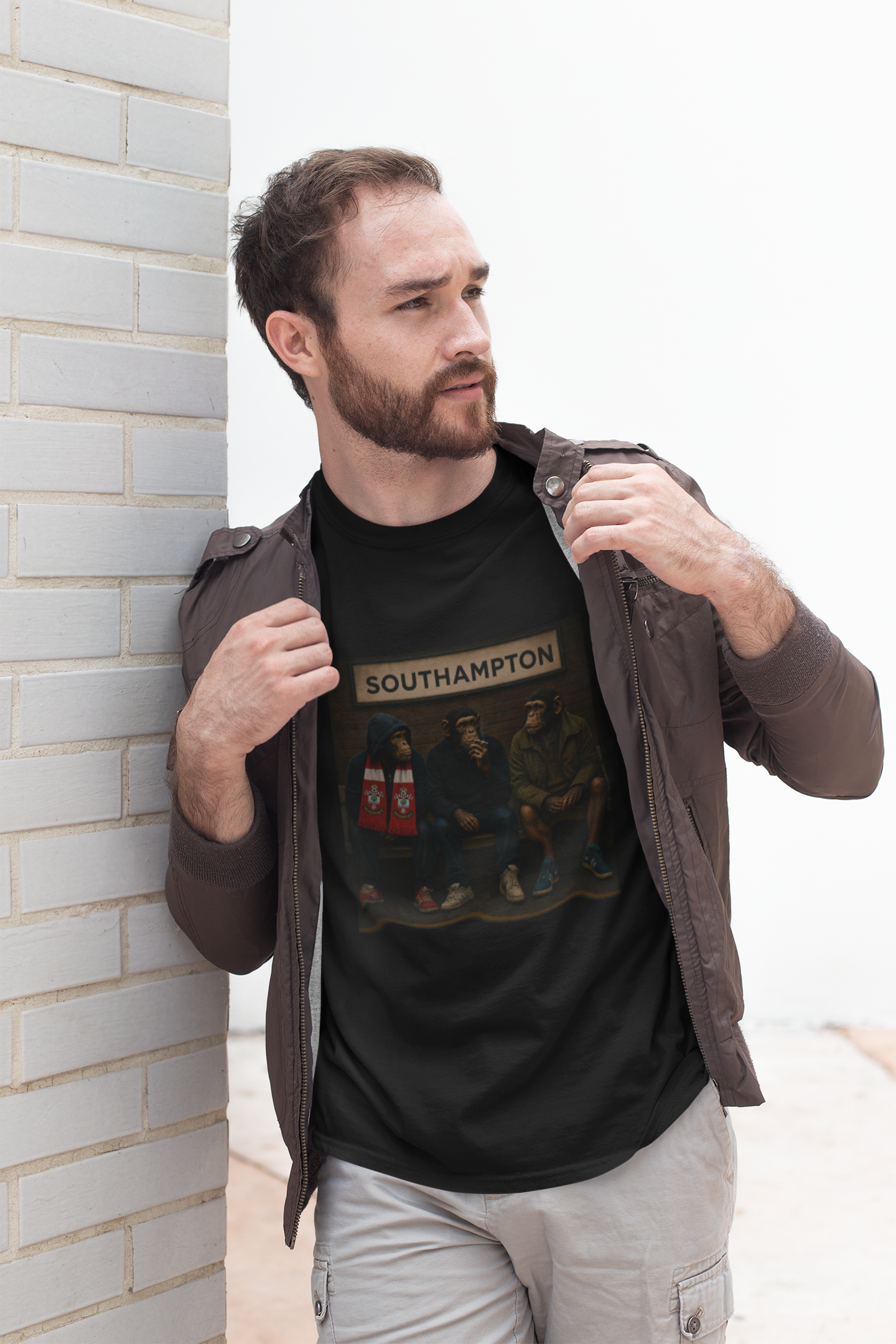A man wearing a black graphic Southampton t-shirt with 'LUTON' printed on the chest, styled with a brown jacket and light gray cargo pants, leaning against a white brick wall.