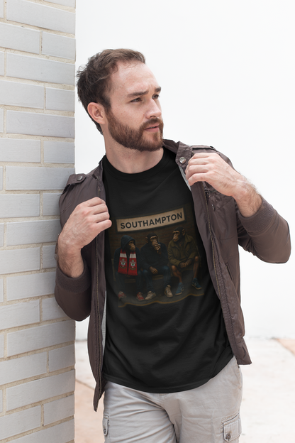 A man wearing a black graphic Southampton t-shirt with 'LUTON' printed on the chest, styled with a brown jacket and light gray cargo pants, leaning against a white brick wall.