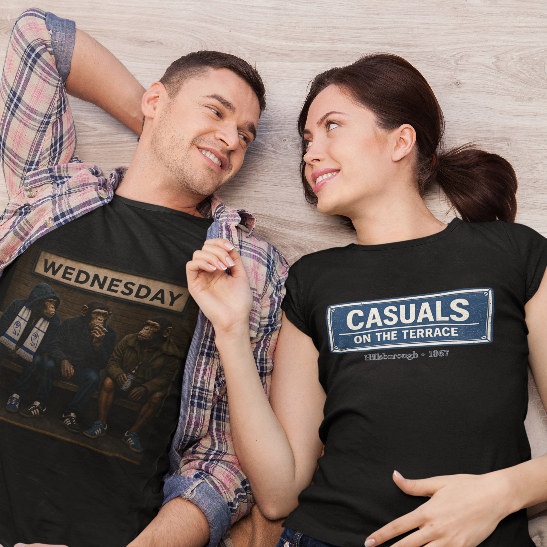 A man and a woman lying on a light wood floor, both wearing black graphic swfc t-shirts. The man's shirt features a 'WEDNESDAY' graphic with a painting of people and a coffee mug, while the woman's shirt features a blue and white street-sign style graphic that reads 'CASUALS ON THE TERRACE' with 'Birmingham - 1867' underneath.