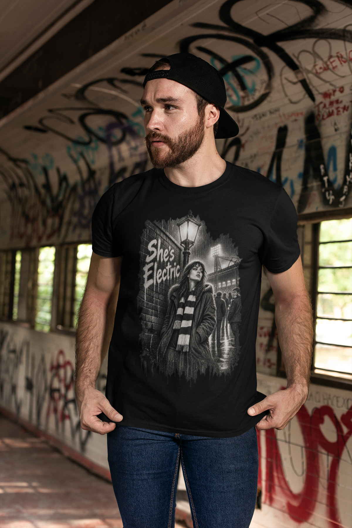 Bearded man in a cap wearing black Oasis She's Electric tee by a graffiti wall