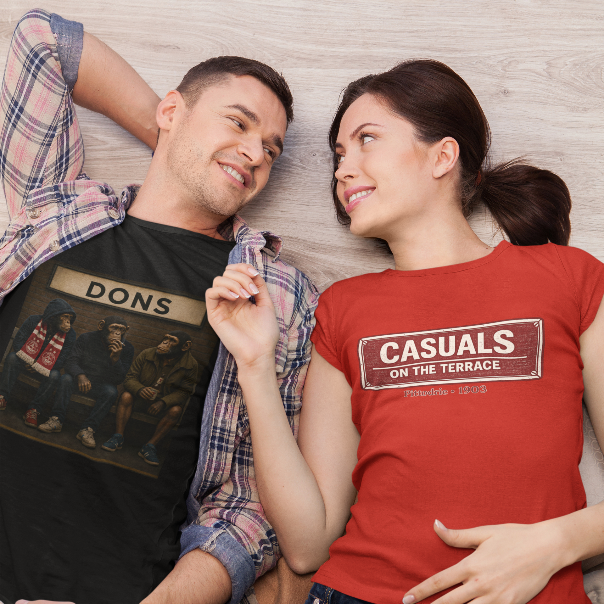 A man and a woman lying on a light wood floor, wearing matching graphic t-shirts. The man wears a black Aberdeen t-shirt with a 'DONS' graphic and an open plaid shirt, while the woman wears a red Aberdeen t-shirt with a 'CASUALS ON THE TERRACE' sign graphic.