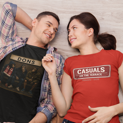 A man and a woman lying on a light wood floor, wearing matching graphic t-shirts. The man wears a black Aberdeen t-shirt with a 'DONS' graphic and an open plaid shirt, while the woman wears a red Aberdeen t-shirt with a 'CASUALS ON THE TERRACE' sign graphic.