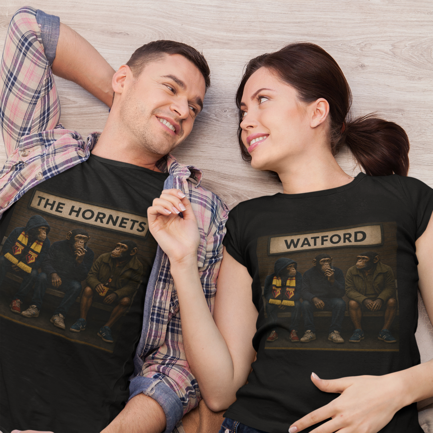 A man and a woman lying side by side on a light wood floor, both wearing matching black graphic WATFORD t-shirts with 'THE HORNETS' and '' printed above a painted illustration of three men in a car.
