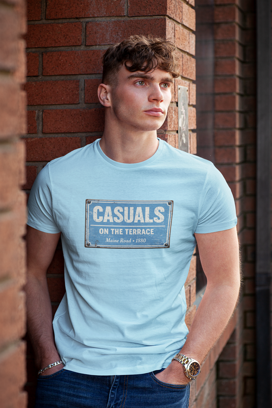 A young man wearing a light blue MCFC t-shirt with a vintage-style blue and white street sign graphic that reads 'CASUALS ON THE TERRACE'. He is leaning against a red brick wall.