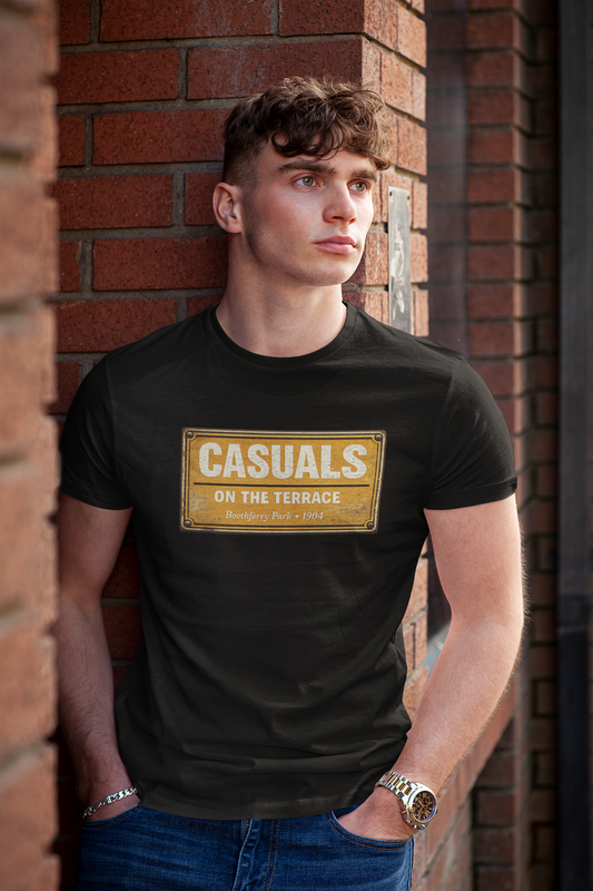 A man wearing a black HULL CITY t-shirt with a vintage-style gold sign graphic that reads 'CASUALS ON THE TERRACE', standing against a red brick wall.