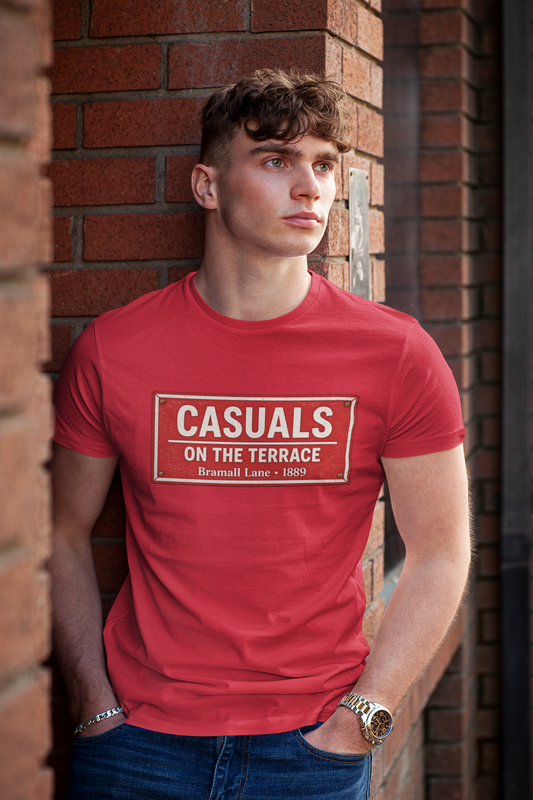 A young man wearing a red SUFC t-shirt with a white graphic print that reads 'CASUALS ON THE TERRACE Bromwell Lane - 1800'. He is leaning against a red brick wall, wearing blue jeans and a gold watch.