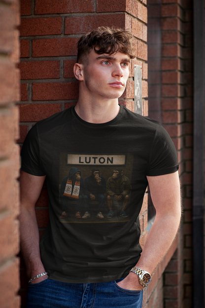 A young man with curly brown hair leans against a red brick wall, wearing a black graphic Luton t-shirt with 'LUTON' printed on the front above a dark illustration, paired with blue jeans.