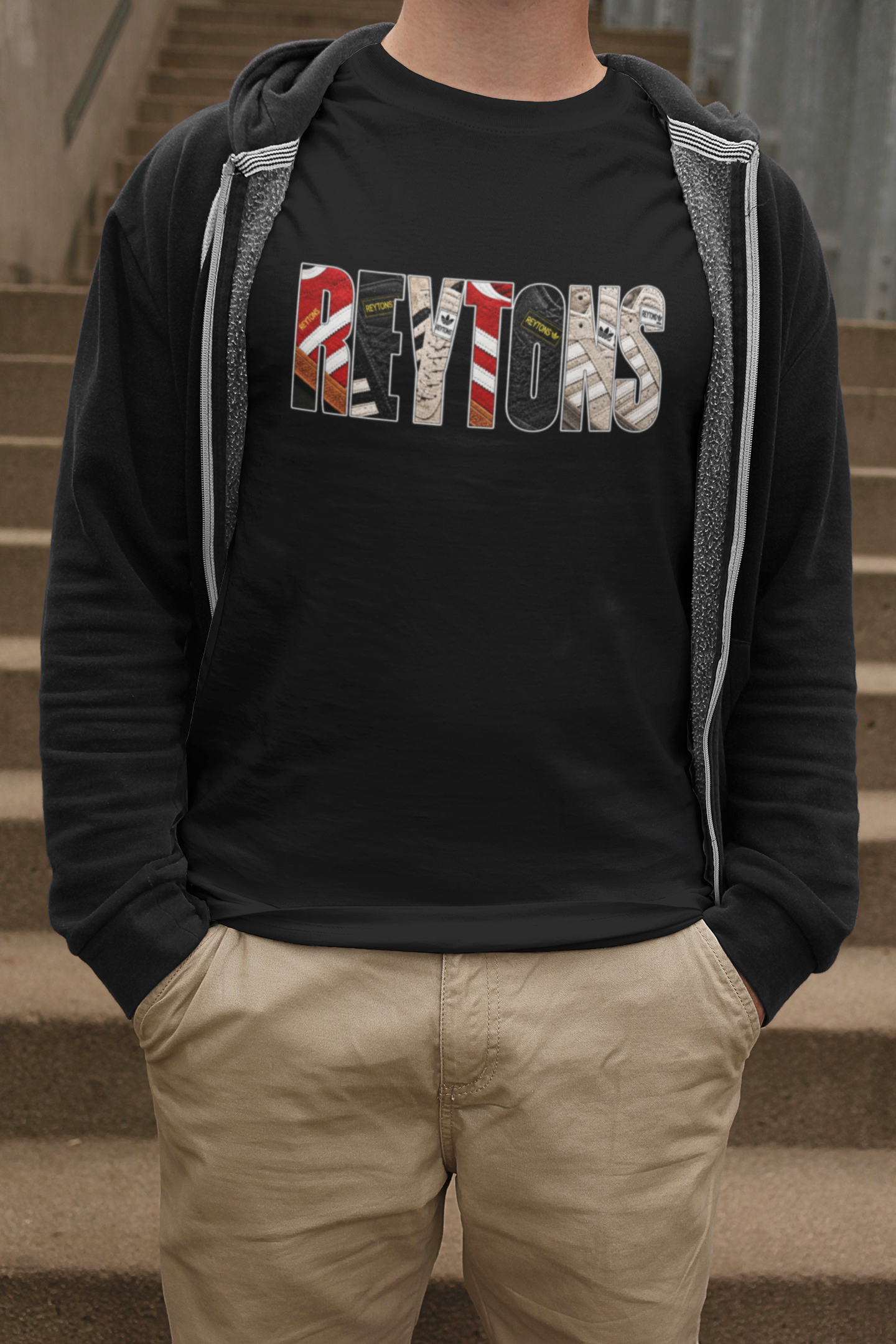 A person wearing a black graphic Reytons t-shirt with a 'Reytons' logo made of road textures, layered under an open black zip-up hoodie with gray lining, paired with khaki pants, standing on concrete stairs.