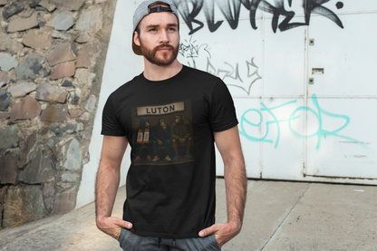 A man wearing a black graphic Luton t-shirt with a 'LUTON' band print, standing outdoors against a white graffiti-covered wall.