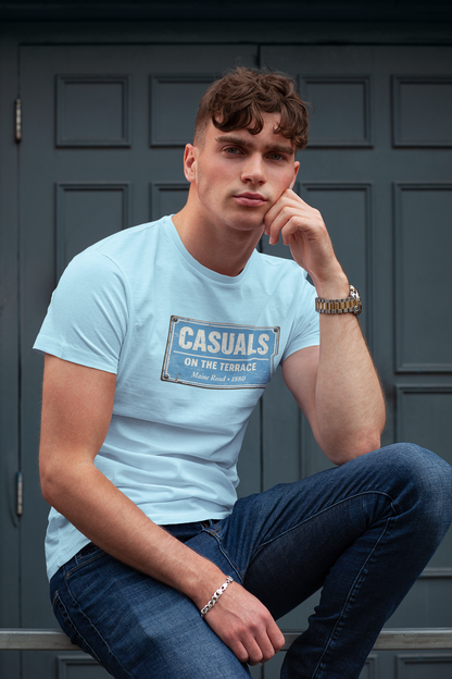 A man wearing a light blue MCFC t-shirt with a vintage-style 'Casuals On The Terrace' sign print, paired with dark blue jeans, sitting in front of a dark gray paneled door.