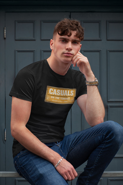 A man wearing a black HULL CITY t-shirt with a gold graphic that reads 'CASUALS ON THE ROAD' and blue jeans, sitting in front of a dark paneled door.