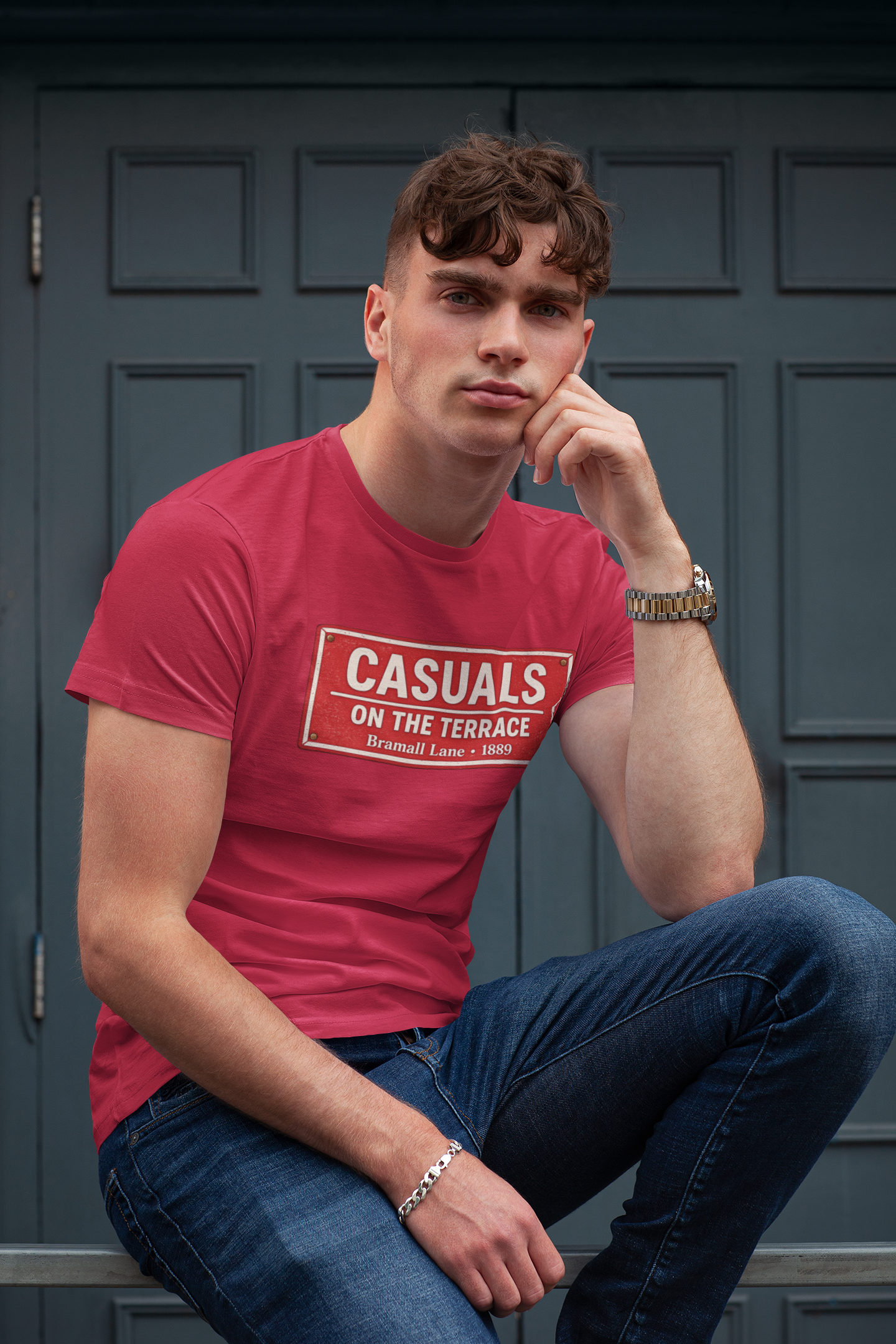 A man wearing a red SUFC t-shirt with a white and black rectangular print that reads 'CASUALS ON THE TERRACE', paired with blue jeans, sitting in front of a dark gray paneled door.