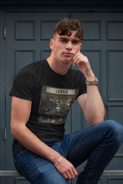 A man wearing a black graphic Leeds t-shirt with 'LEEDS' printed on the front, paired with blue jeans, posing in front of a dark paneled door.