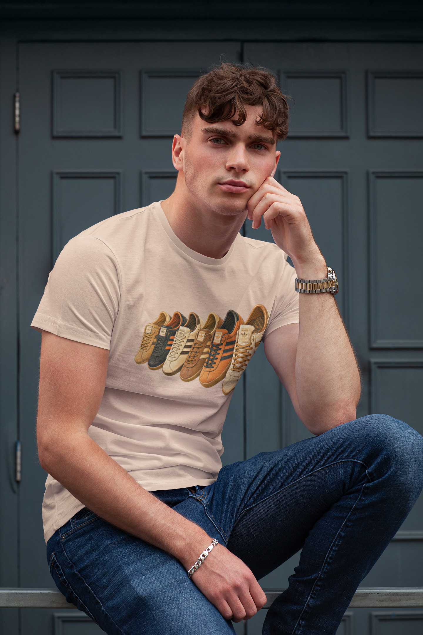 A man wearing a beige graphic t-shirt with a sneaker print, blue jeans, and metal bracelets, sitting in front of a dark blue paneled door.