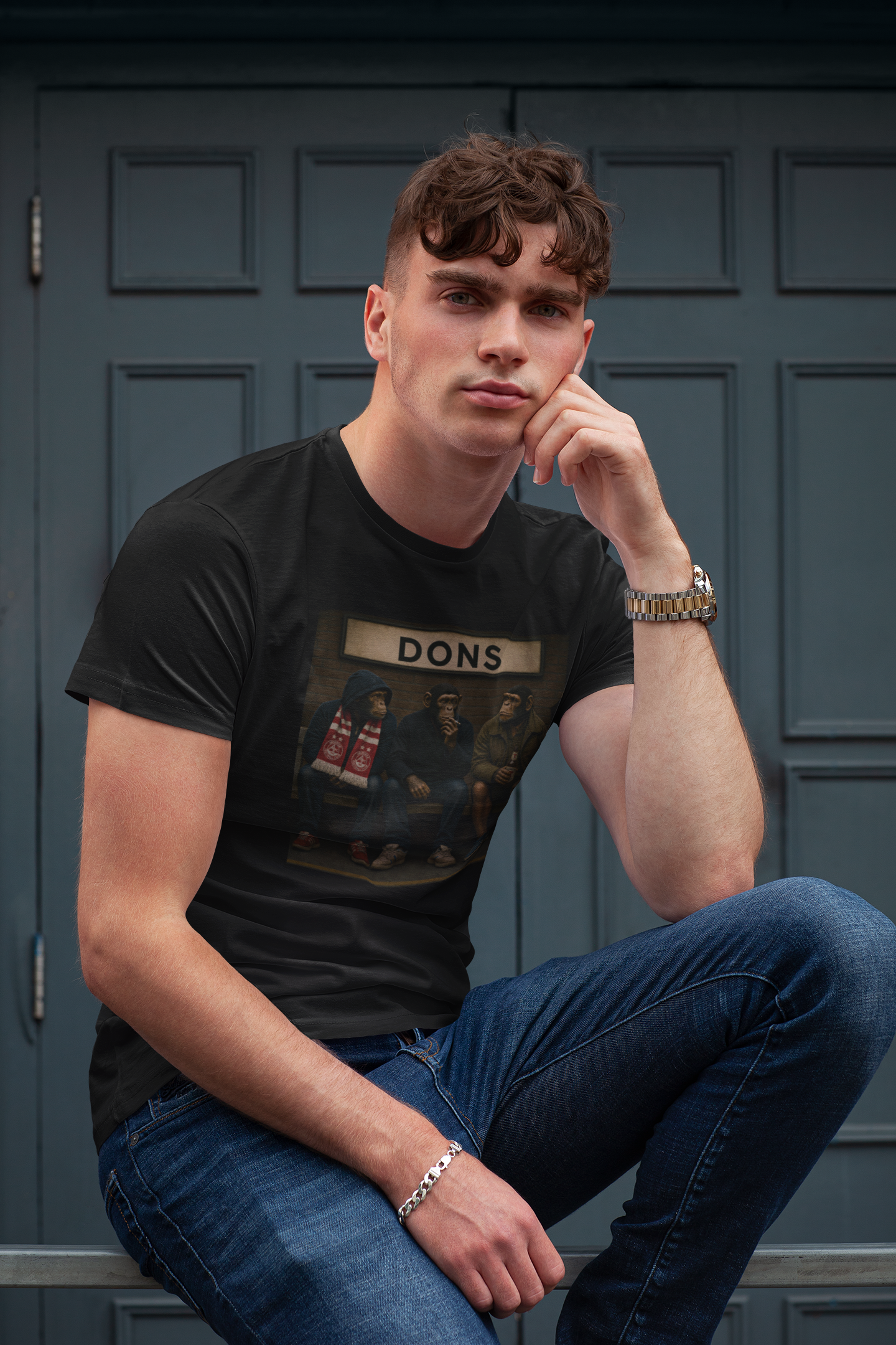 A man wearing a black graphic Aberdeen t-shirt with a Aberdeen print, paired with blue jeans, posing in front of a dark paneled door.
