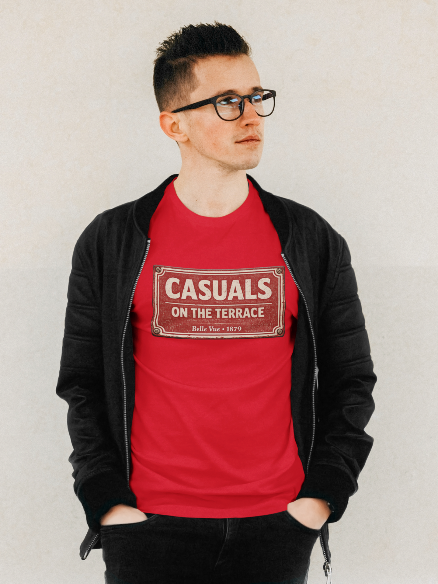 A man wearing a red DRFC t-shirt flat lay featuring a central vintage red and white street sign graphic reading "CASUALS ON THE TERRACE" with "Belle Vue • 1879" printed below, and an inner neck label for On The Terrace., layered under a black zip-up jacket and wearing black-rimmed glasses, standing against a light textured wall.