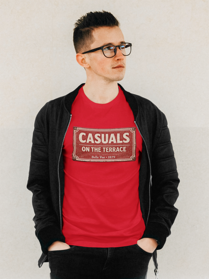 A man wearing a red DRFC t-shirt flat lay featuring a central vintage red and white street sign graphic reading "CASUALS ON THE TERRACE" with "Belle Vue • 1879" printed below, and an inner neck label for On The Terrace., layered under a black zip-up jacket and wearing black-rimmed glasses, standing against a light textured wall.